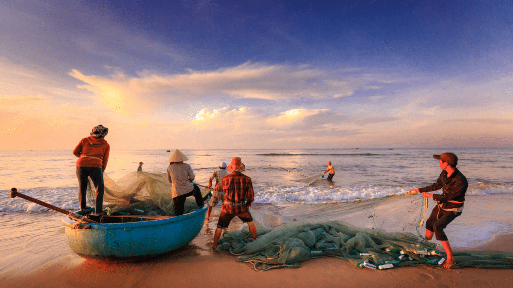 a person with a hat walking on the beach with baskets of fish